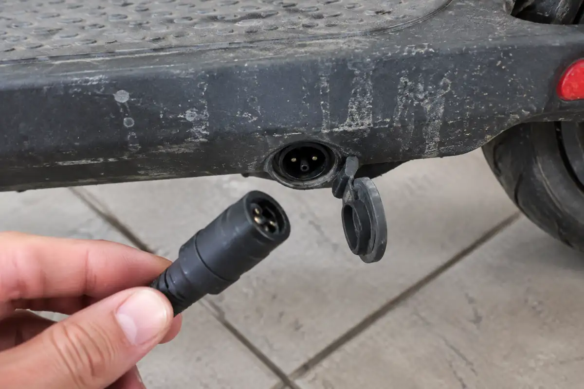 Prise de charge au bas du plateau d'une trottinette électrique, capuchon ouvert, avec la fiche du chargeur tenue devant